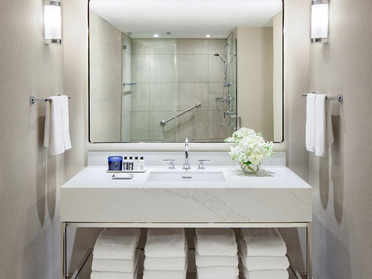 This image shows a modern bathroom with a large mirror, sink, towels on racks, vanity with folded towels, flowers on counter, and a glass-enclosed shower.