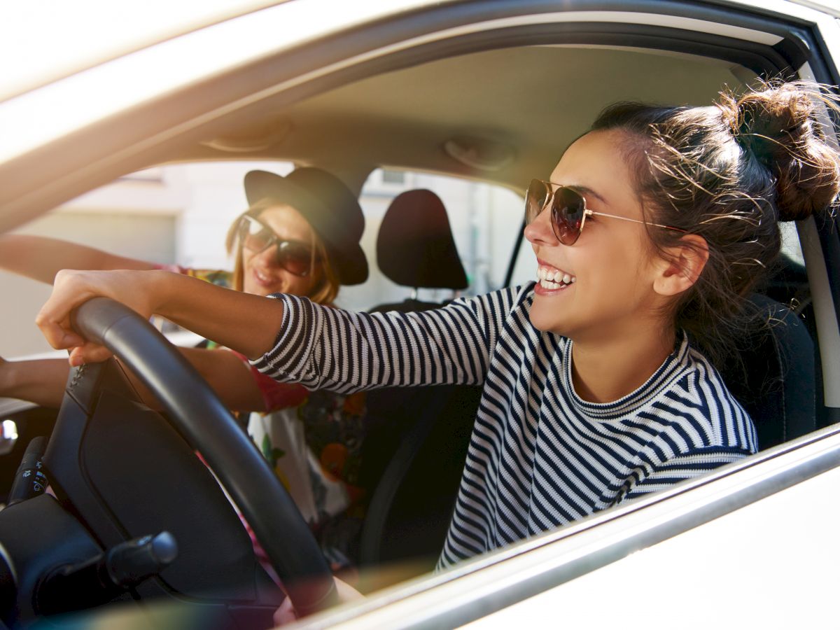 Two people are sitting in a car; the driver is laughing and wearing sunglasses and a striped shirt. The passenger also wears sunglasses and a hat.