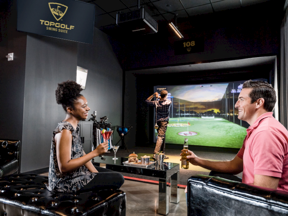 Three people are at a golf simulation lounge with drinks, and one person is taking a swing in front of a virtual golf screen.