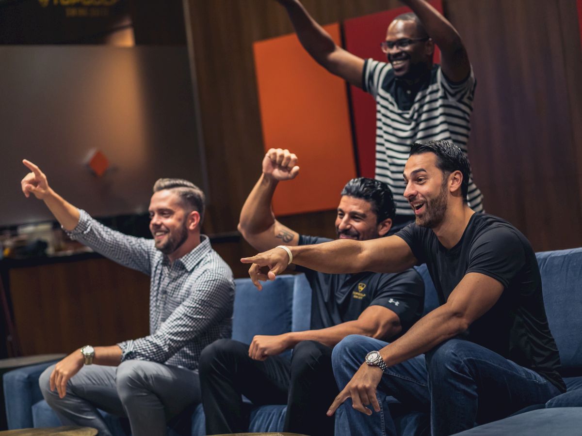 A group of four friends excitedly pointing at something off-camera, possibly a TV, with one standing and the others sitting on a couch.