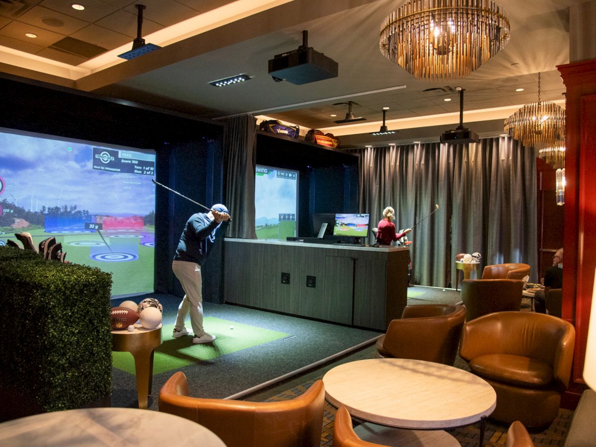 The image shows an indoor golf simulator setup in a modern lounge area with people playing virtual golf.