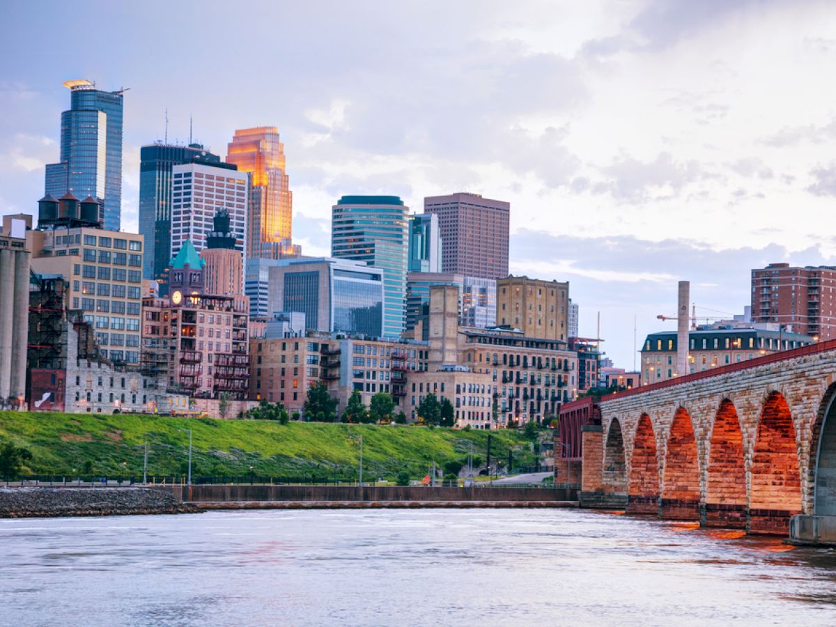 The image shows a cityscape with modern skyscrapers and an arched stone bridge over a river, likely at sunset, casting a warm glow over the buildings.