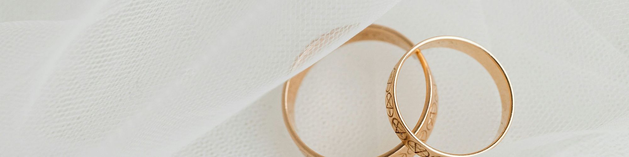 Two gold wedding rings with intricate designs are placed on a white, textured fabric.