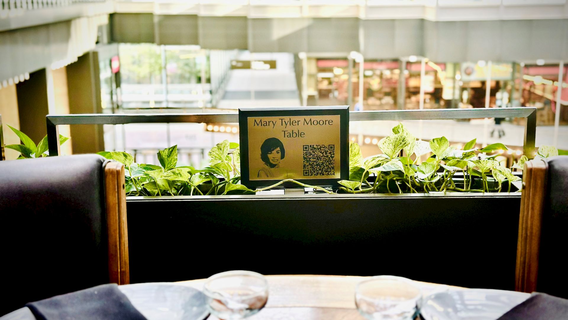 The image shows a restaurant table set for two with a sign labeled "Mary Tyler Moore Table" and a QR code in an indoor setting.