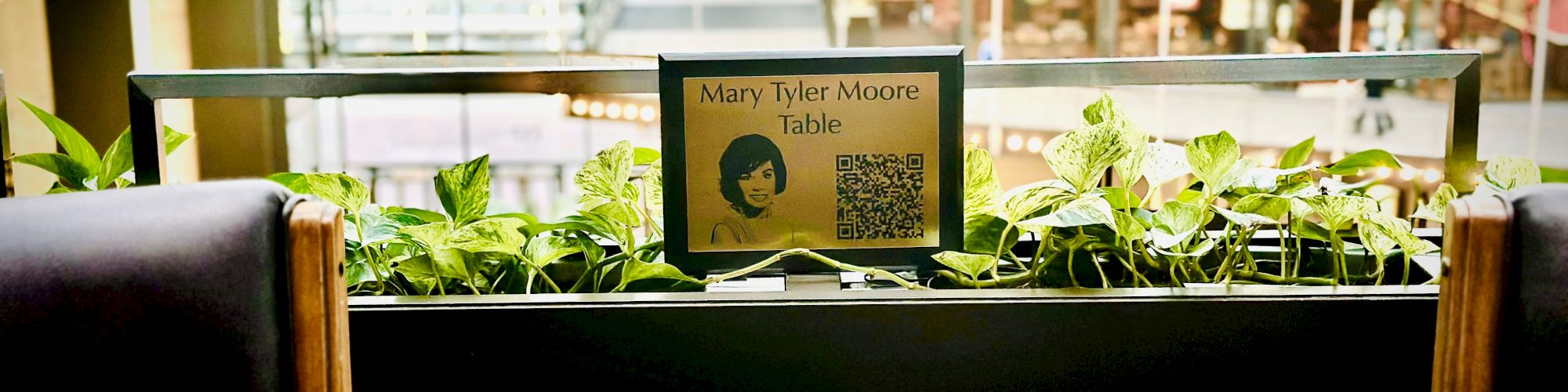 The image shows a restaurant table set for two with a sign labeled "Mary Tyler Moore Table" and a QR code in an indoor setting.