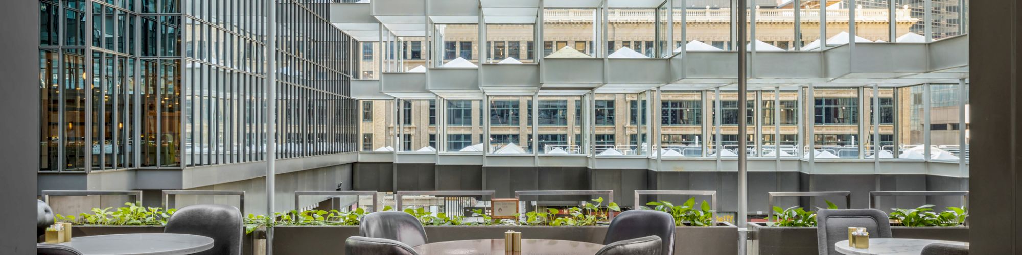 An outdoor seating area with round tables and chairs, surrounded by modern architecture and greenery, with a view of city buildings in the background.