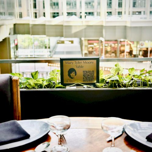 A dining table set for two with a sign that reads "Mary Tyler Moore Table" and a QR code, set against a backdrop of a building interior.