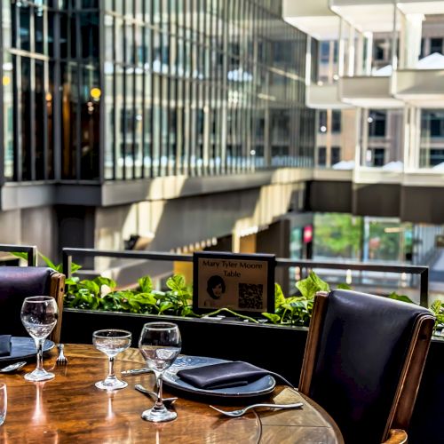 A sunlit restaurant table set with glasses and napkins, overlooking a cityscape with modern buildings in the background.