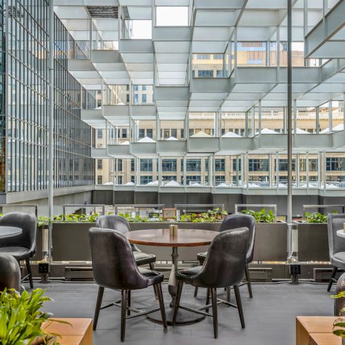 An outdoor seating area with round tables and chairs, surrounded by a modern, geometric pergola and planters, set against an urban backdrop.