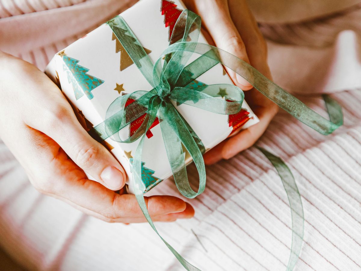 A person holds a small gift wrapped in festive paper with Christmas tree patterns, tied with a green ribbon.