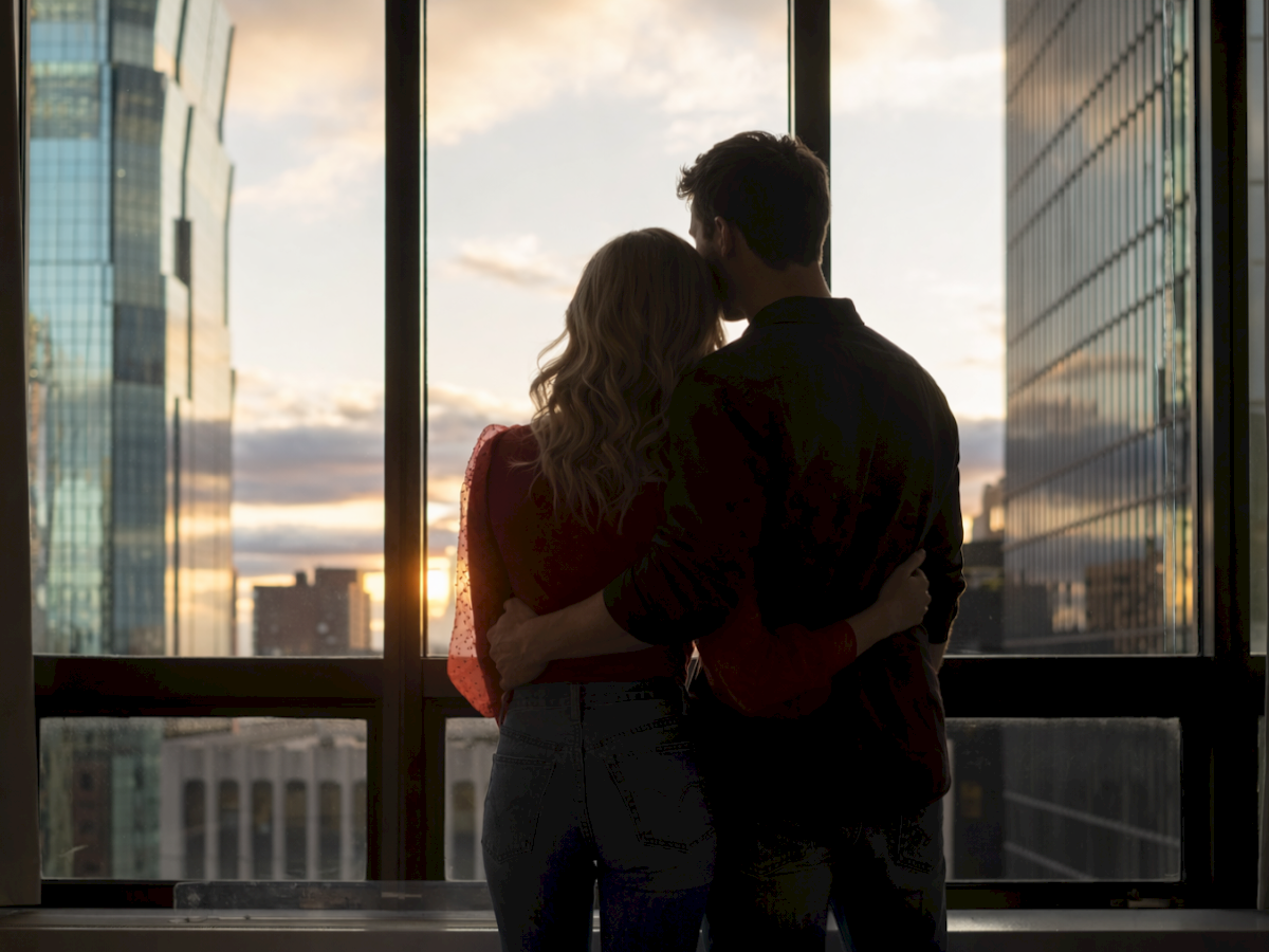 A couple stands close, silhouetted against a city sunset, embracing as they look out a large window between tall buildings.