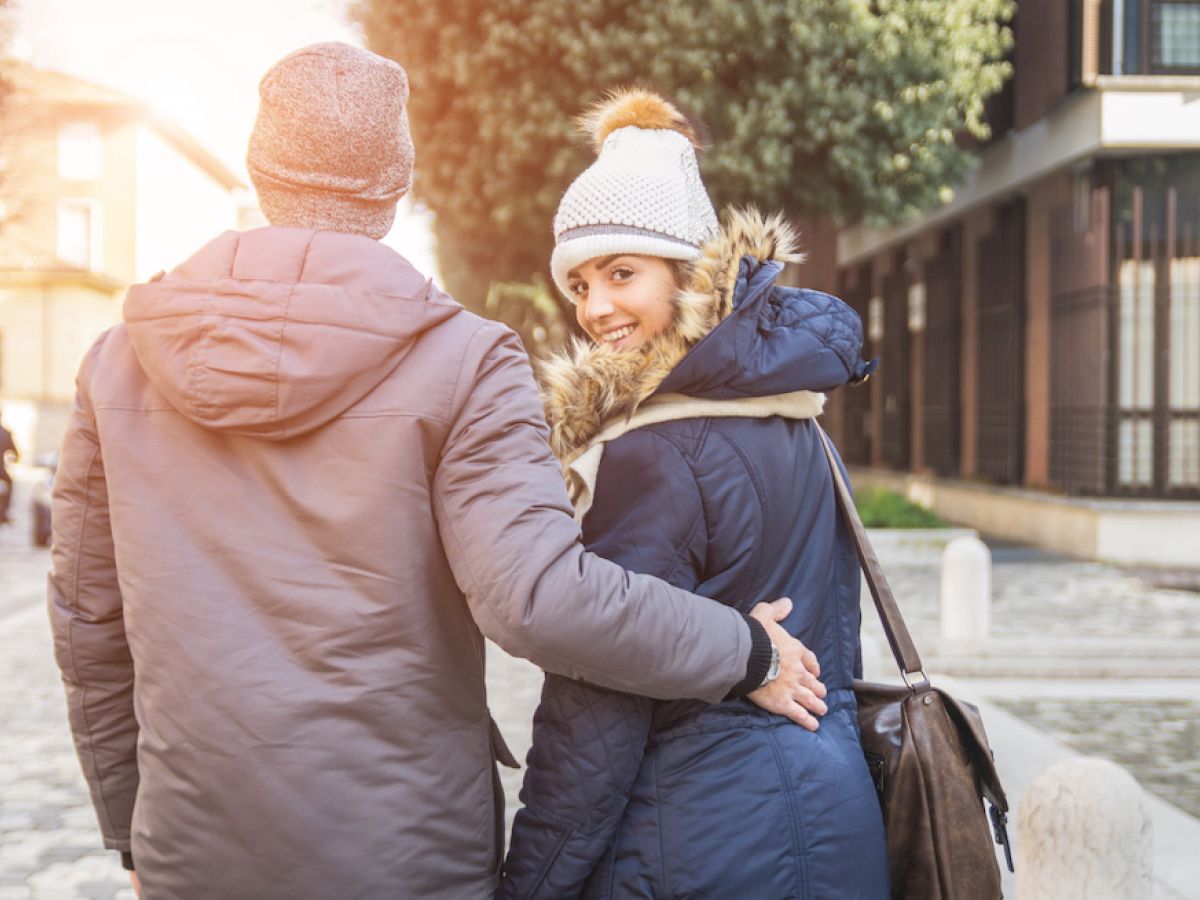 A couple dressed warmly in winter clothing is walking outdoors, with the woman smiling and looking back at the camera.