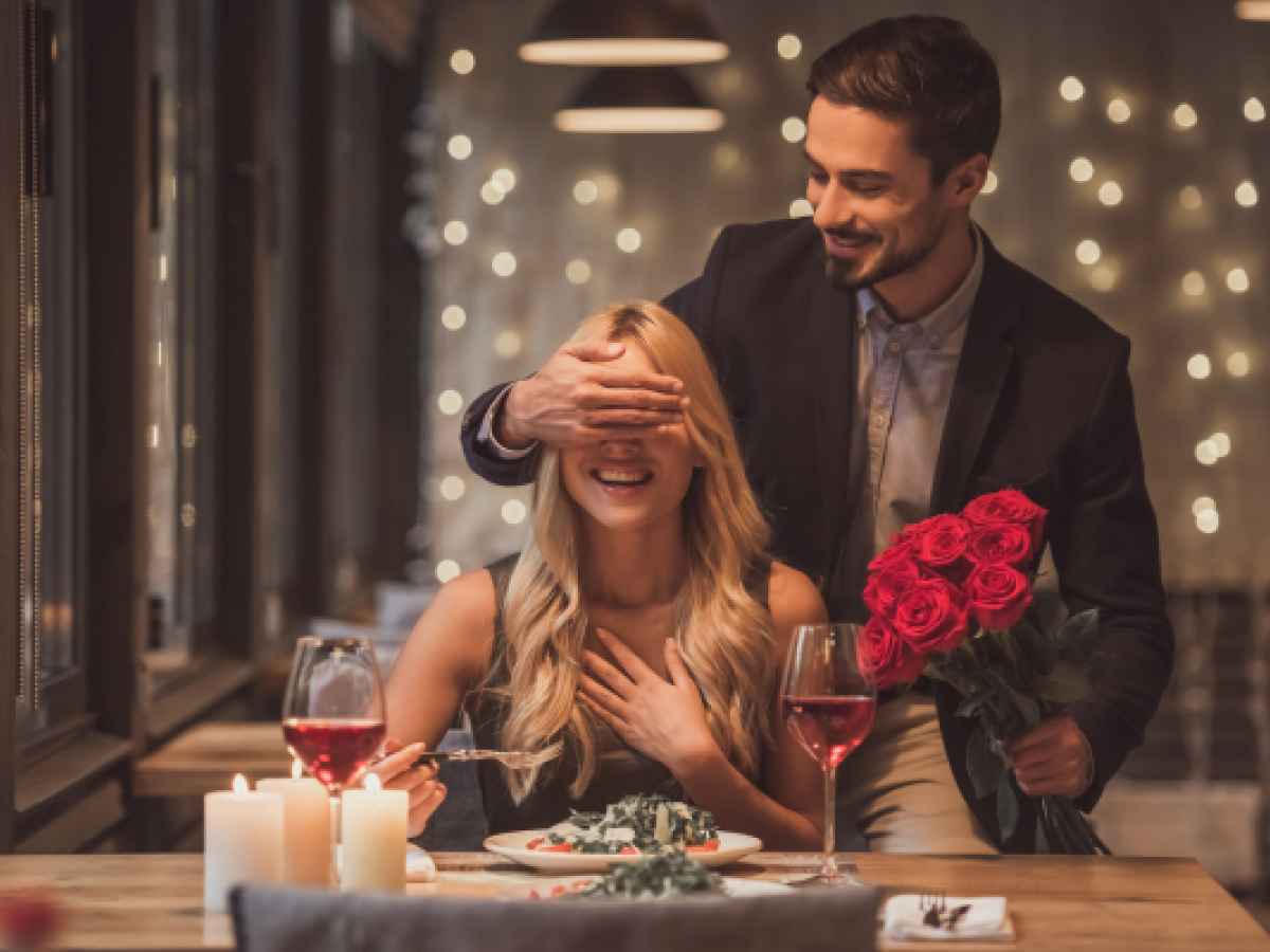 A man surprises a woman at a candlelit dinner, holding red roses while covering her eyes, creating a romantic atmosphere.