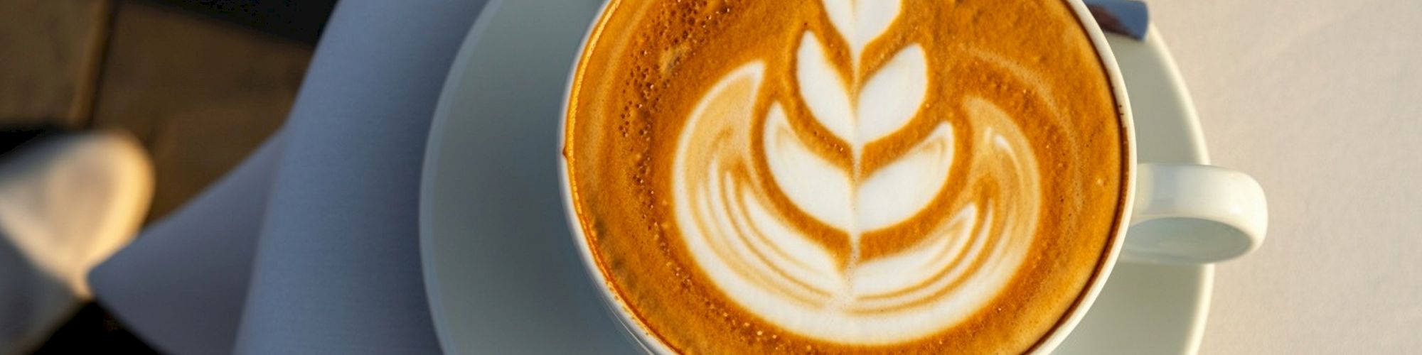 A cup of cappuccino with latte art in a leaf pattern on a saucer, placed on a white tablecloth in soft lighting.