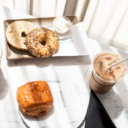 Bagels with cream cheese, a pastry, and two coffee cups are on a table in sunlight.