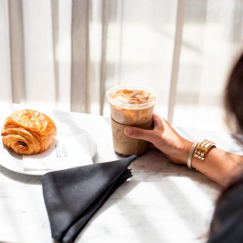A person holds an iced coffee next to a croissant on a white plate, with a black napkin on a marble table.