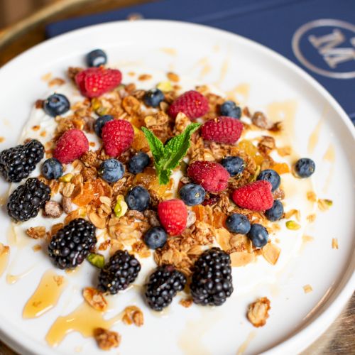 A bowl of yogurt topped with granola, raspberries, blackberries, blueberries, mint, and honey drizzle on a white plate.
