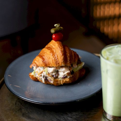 A croissant sandwich with a skewer and a cherry tomato, served with a glass of iced matcha on a table.