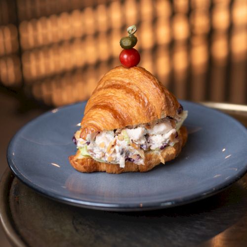 A croissant filled with chicken salad is on a blue plate, topped with an olive and cherry tomato skewer.