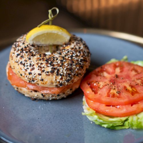 A bagel sandwich with seeds and a lemon slice on top, filled with salmon. Lettuce and tomato slices are on the side, on a blue plate.