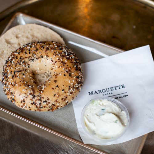 A bagel with seeds, cream cheese in a cup, and a "Marquette" napkin are on a tray.