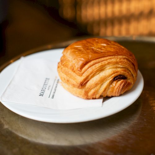 A chocolate croissant on a white plate with a napkin underneath, placed on a round metal table.