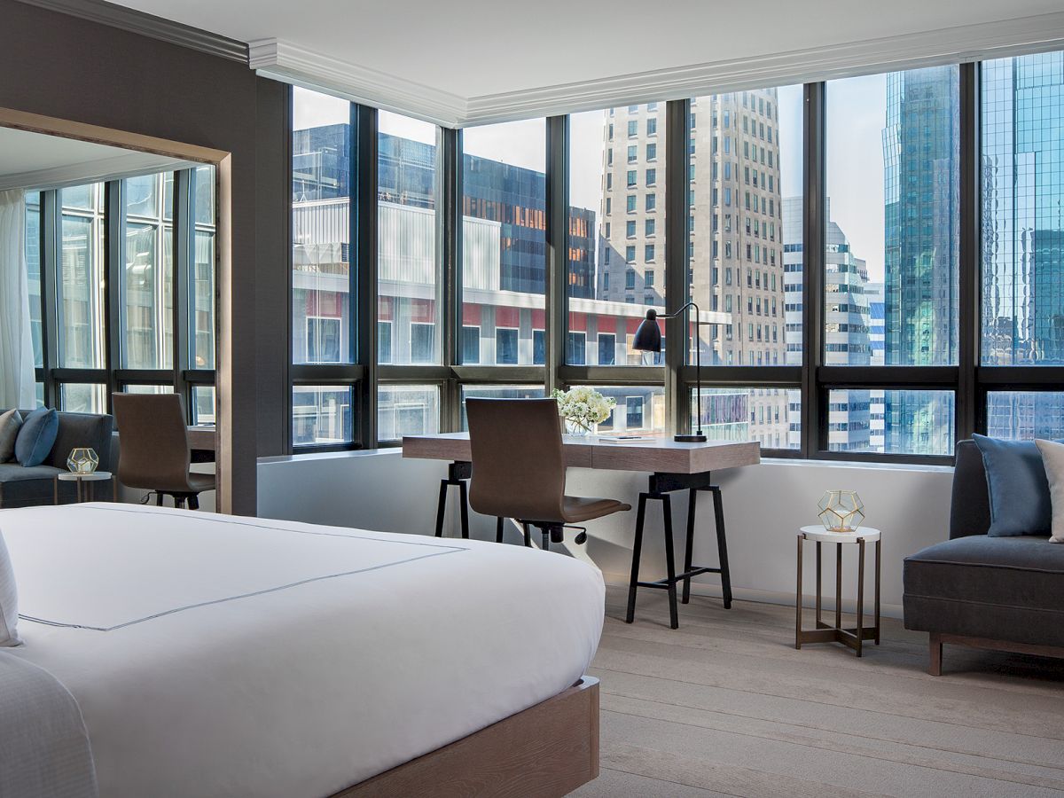 A modern hotel room with a bed, desk, chair, couch, and large windows showing a city skyline.
