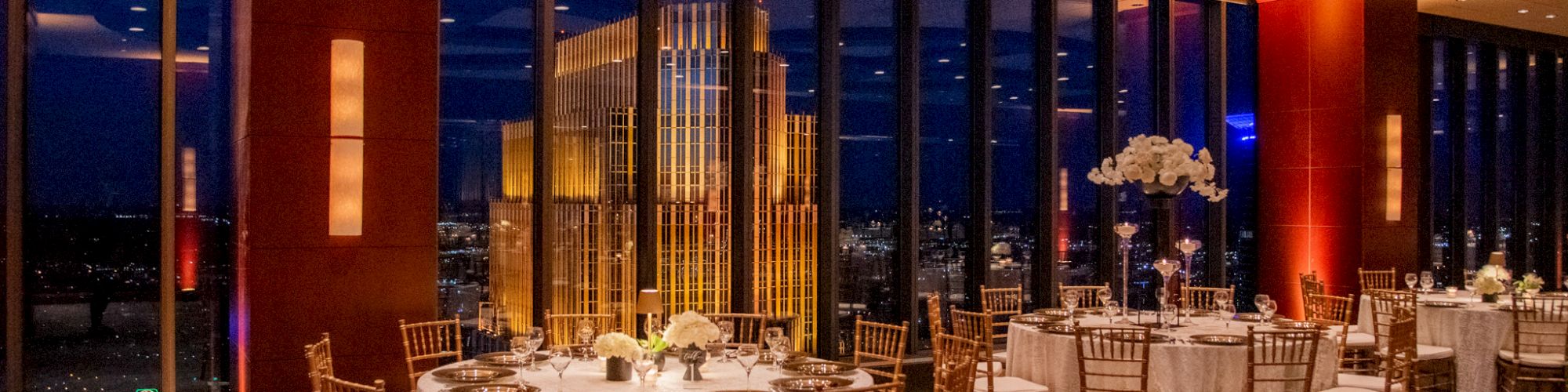 Elegant dining area with round tables, Chiavari chairs, and cityscape view from large windows. Evening ambiance with soft lighting.