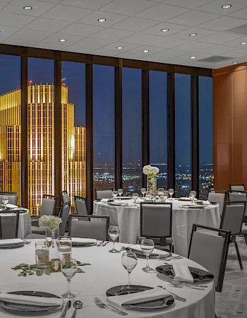 Elegantly set dining tables in a room with large windows, overlooking a beautifully illuminated skyscraper and city lights at night.