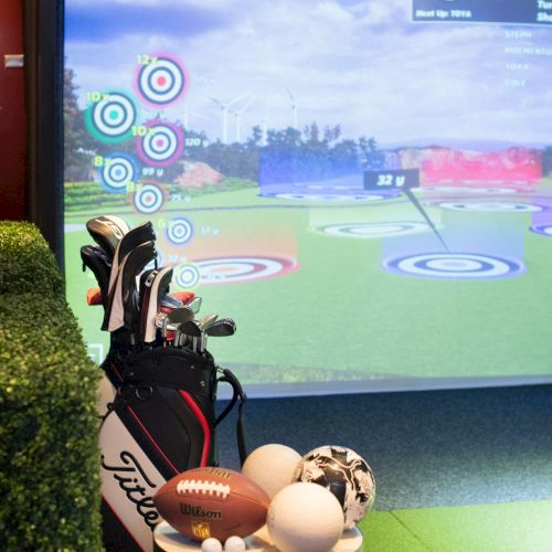 The image shows a golf simulator set up with golf clubs and various sports balls, including a football and soccer ball, in an indoor setting.