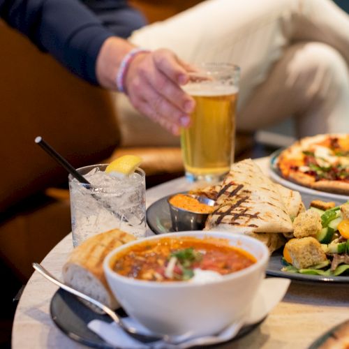 A table with pizza, salad, soup, drinks, and a person holding a glass of beer, all set in a casual dining setting.