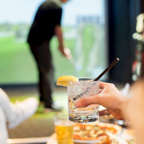 A person holding a drink with a lemon, straw, and ice. In the background, someone plays golf indoors, next to a table with food and drinks.
