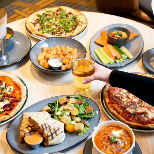 The image shows a table with pizzas, fries, sandwiches, salad, dips, and drinks. A person is reaching for a drink.