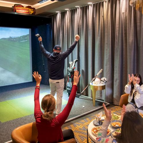Indoor golf simulation with a person celebrating and a group of people enjoying food, raising their hands in excitement.