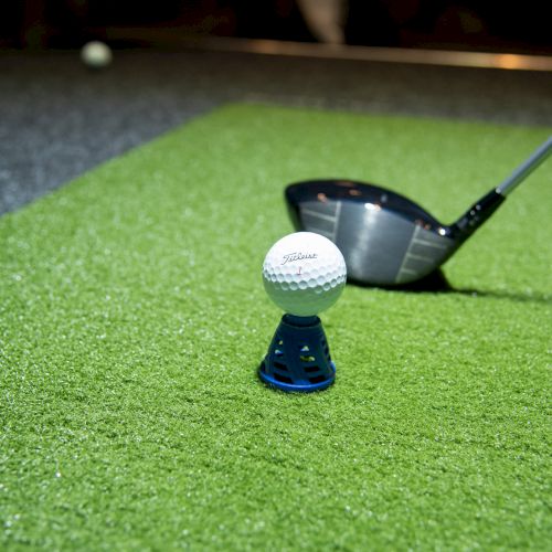 A golf ball on a tee is positioned on a green mat, ready to be hit by a golf club in the image.