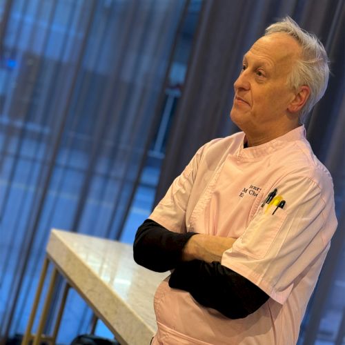 An older man in a pink chef's jacket stands with his arms crossed in front of a window.