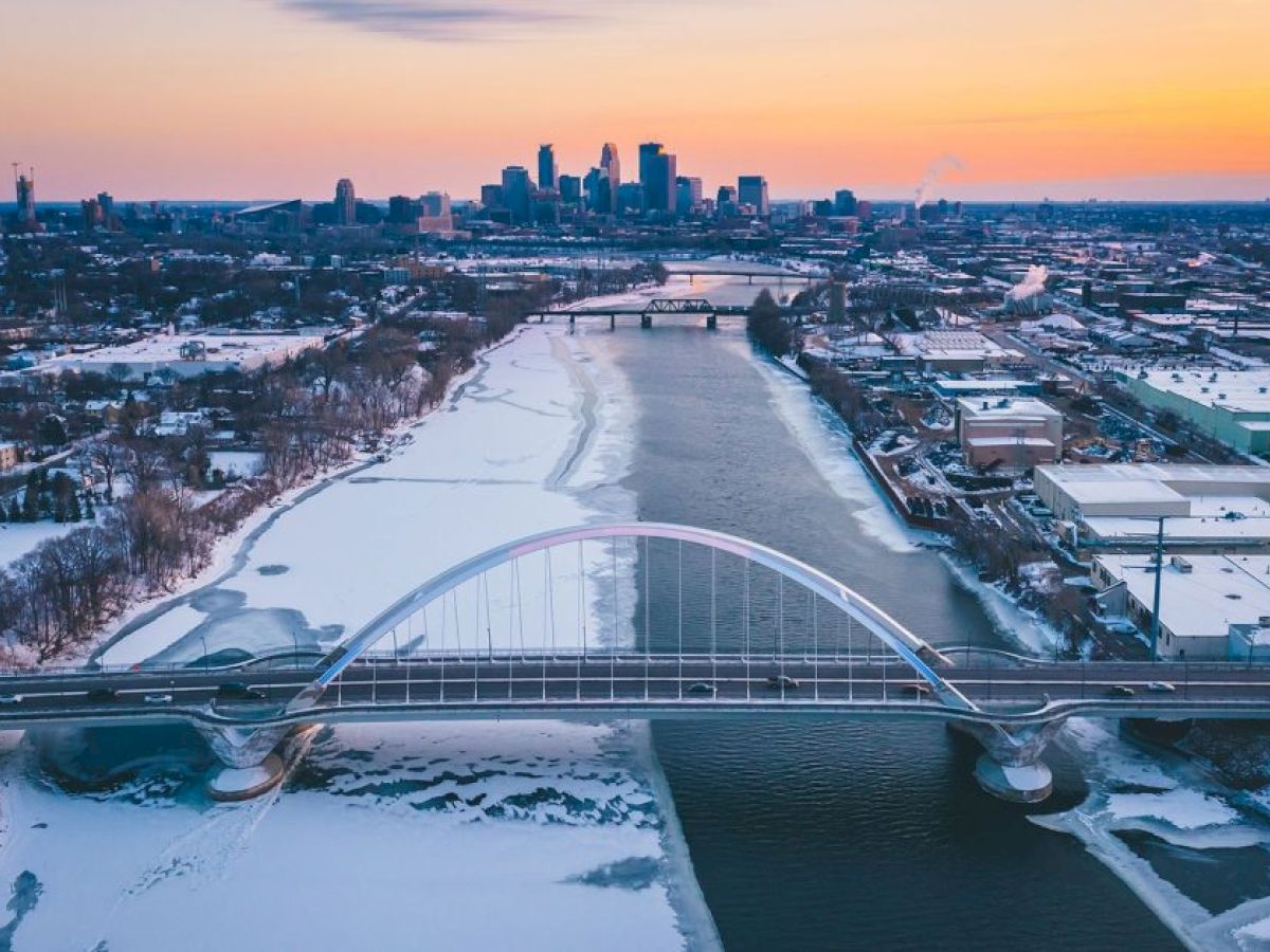 A snowy urban landscape with a bridge spanning a frozen river, city skyline in the background under a colorful sunset.
