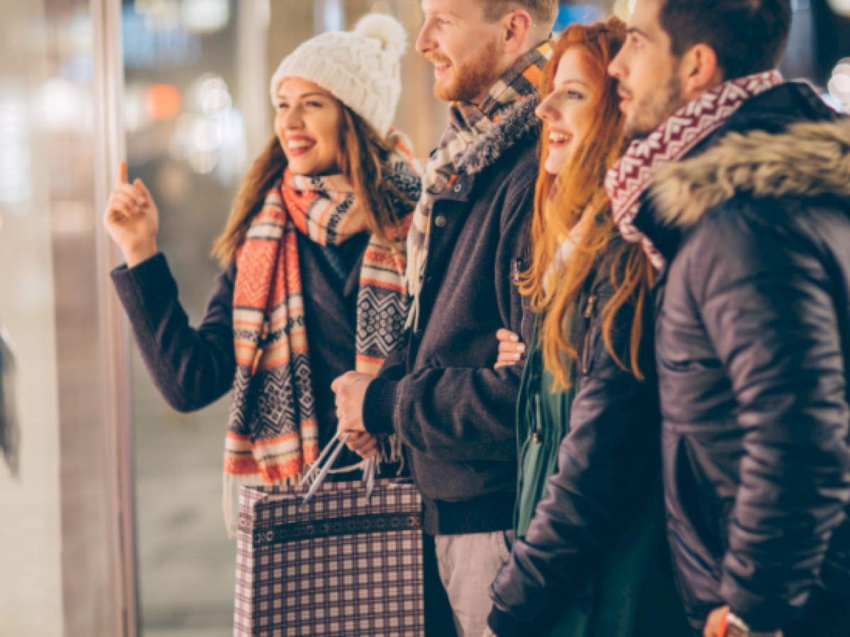 A group of four friends dressed in winter coats and scarves, chatting and smiling as they look at a storefront window.