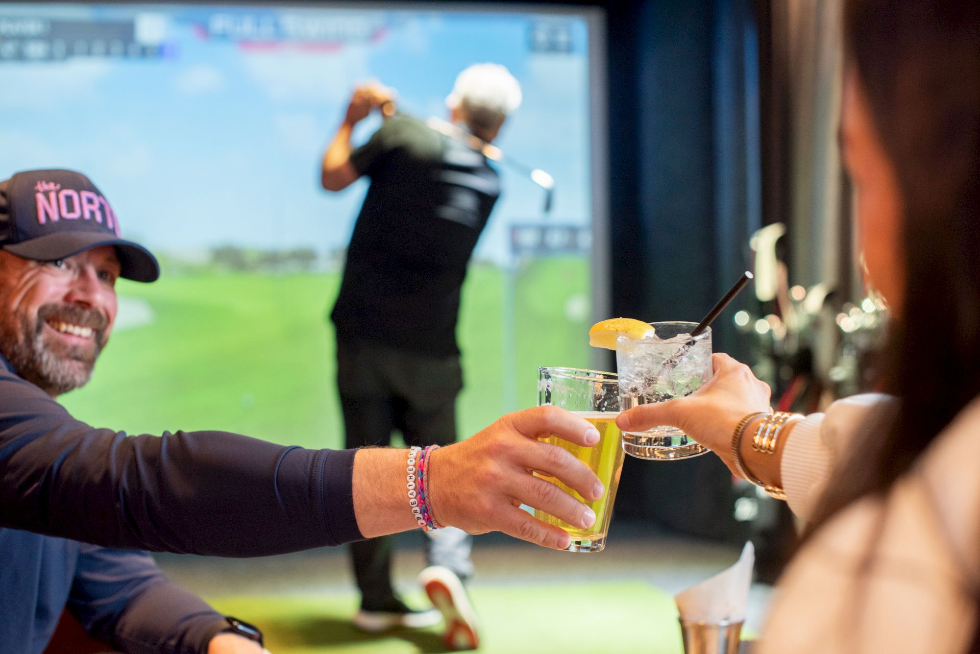 Two friends toast with beers in a golf simulator bar, while a golfer swings in the background.