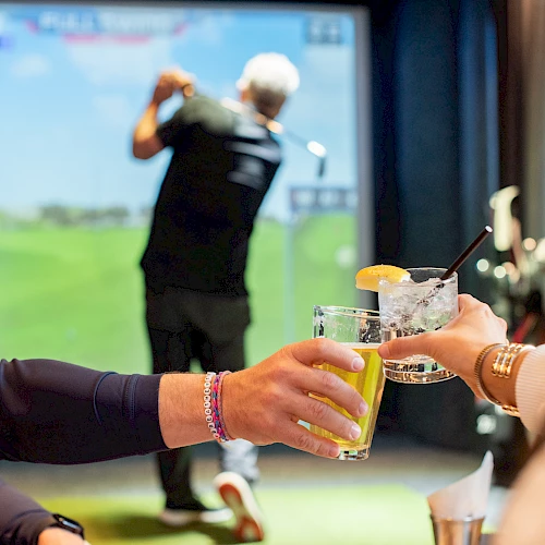 Two people clink beer glasses in a golf simulator room while another person toasts with a drink, golfer in the background.
