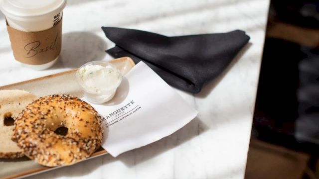A coffee cup, a sesame bagel with cream cheese on a plate, a napkin, and a white cup on a marble table.