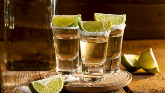 Shot of clear shots with lime wedges on the rim, arranged on a tray and table, suggesting a tequila or citrus liquor setup, rustic vibe.