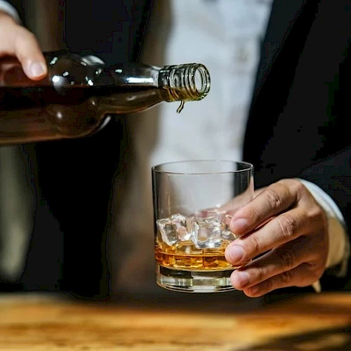 A person in a suit pours whiskey from a bottle into a short glass with ice, standing at a wooden bar.