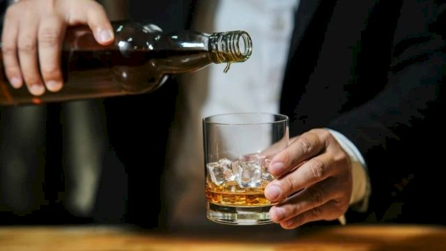 A person in a suit pours whiskey from a bottle into a short glass with ice, standing at a wooden bar.