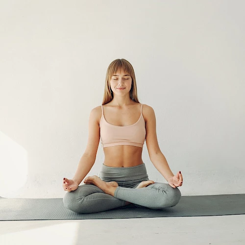A woman in a pink sports bra and gray leggings sits in a cross-legged yoga pose on a mat, meditating in a bright, minimal room.