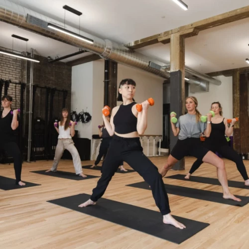 People in a yoga studio lifting small orange dumbbells in a wide-legged stance during a group workout, led by an instructor at the front.