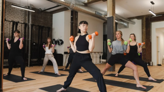 People in a yoga studio lifting small orange dumbbells in a wide-legged stance during a group workout, led by an instructor at the front.
