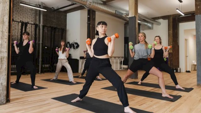 People in a modern gym class doing yoga or martial arts with orange dumbbells, synchronized on mats, focused and balanced, energetic workout.