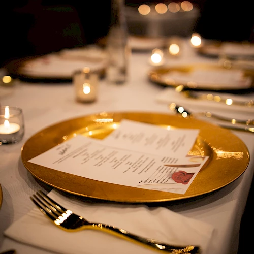 A fancy table setting with a gold charger plate, menu card, candles, and polished silverware in a dimly lit dining room, elegant dinner scene.