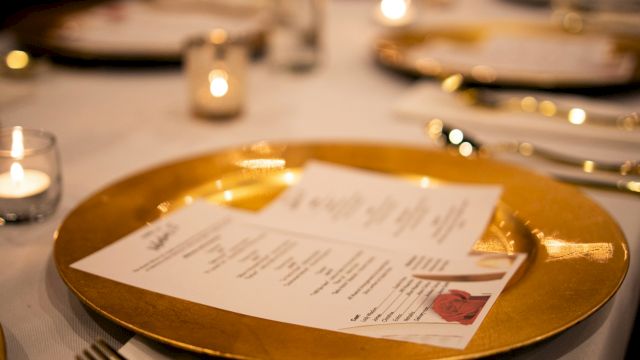 A fancy table setting with a gold charger plate, menu card, candles, and polished silverware in a dimly lit dining room, elegant dinner scene.
